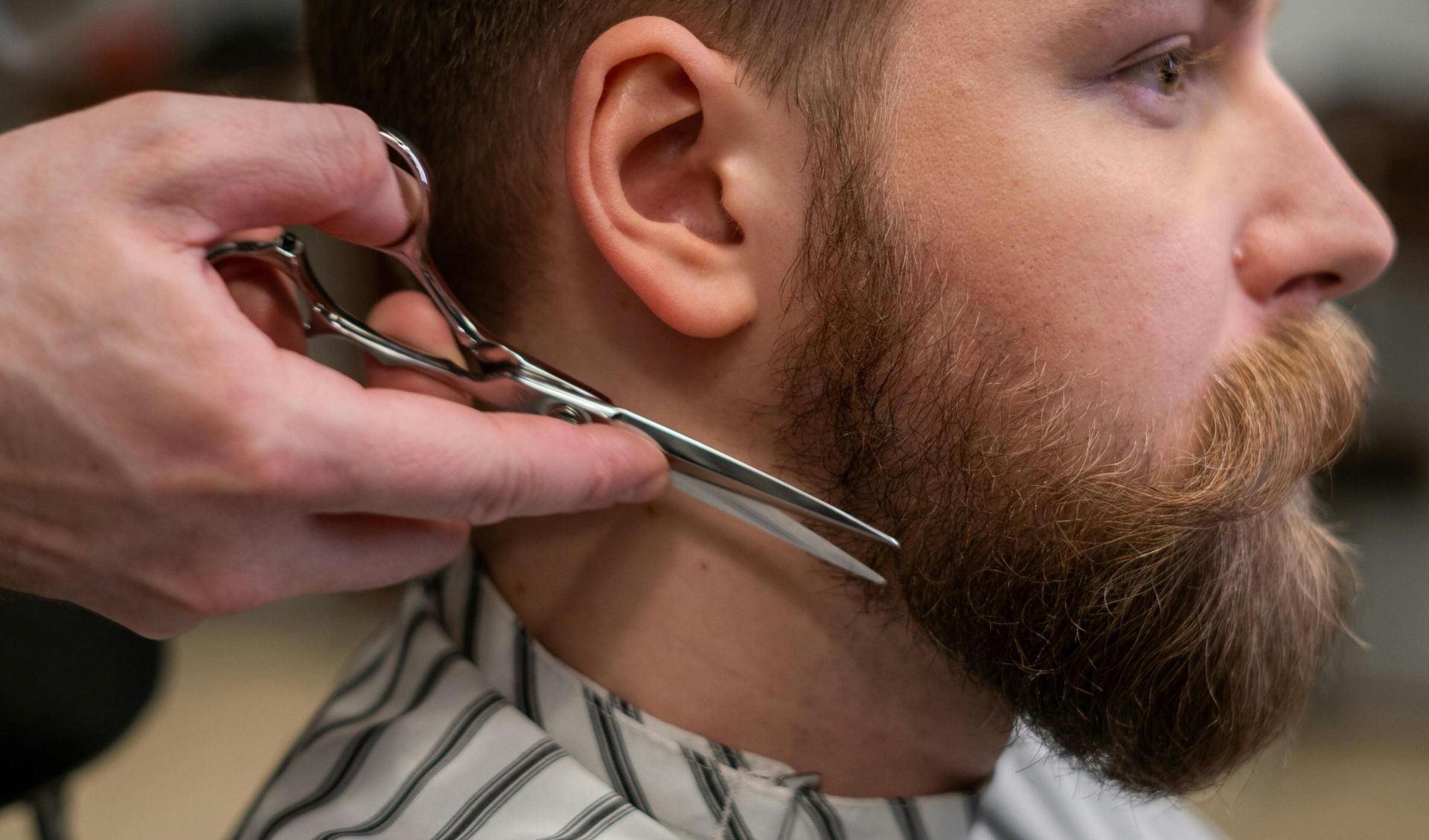 A side view of a barber trimming a bearded man's hair in a barber shop.