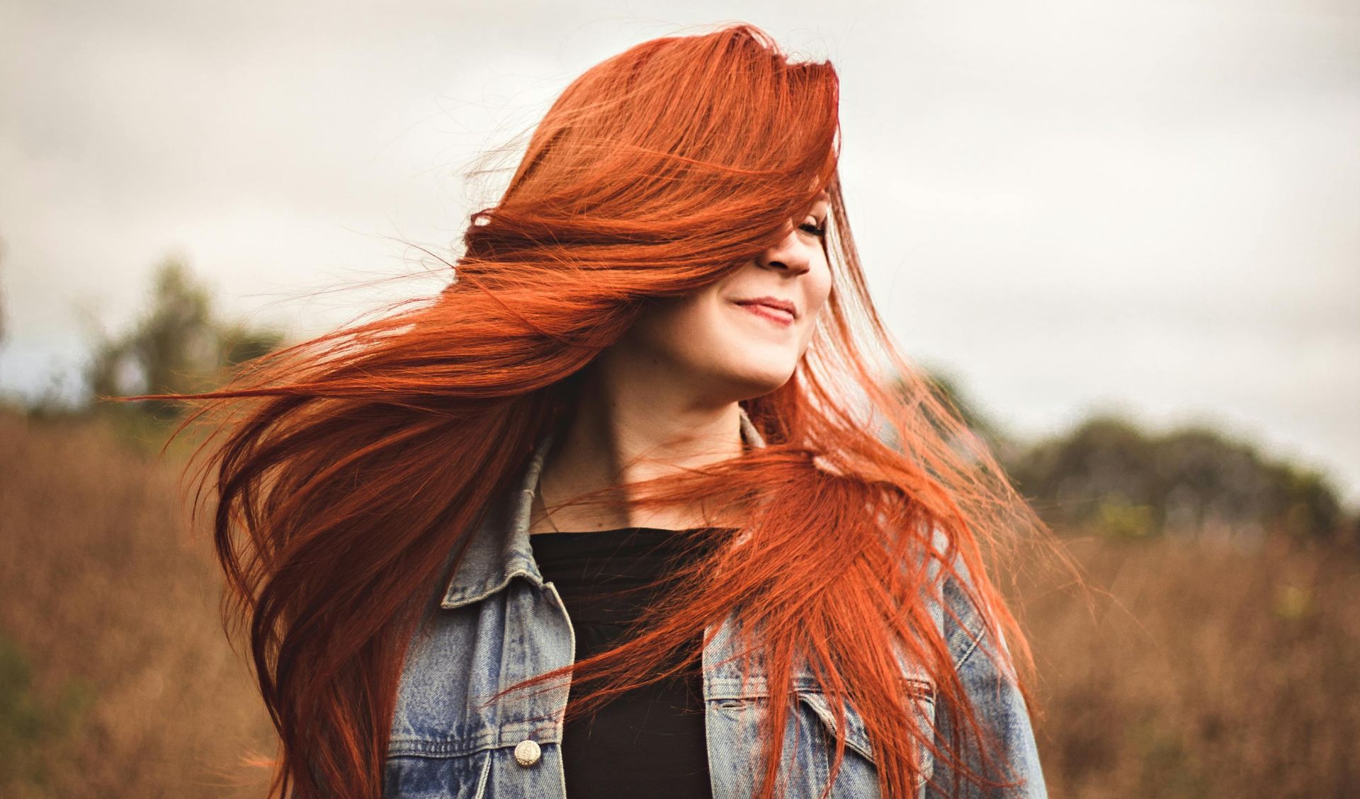 Smiling woman with red hair enjoys a breezy day in a scenic autumn field.